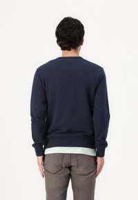 Felpa blu navy con maniche lunghe e scollatura rotonda, realizzata in tessuto morbido. Indossata sopra una maglietta verde chiaro e abbinata a pantaloni grigi.