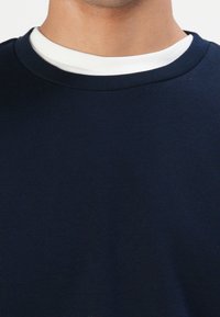 Persona con camisetas de cuello redondo en capas, azul marino y blanca, mostrada desde el cuello hasta el pecho.
