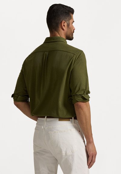 Camisa verde oliva com mangas arregaçadas, colarinho clássico e detalhe de pregas nas costas, combinada com calças brancas com bolsos traseiros.