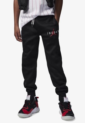 Parte inferior del cuerpo con pantalones deportivos Jordan negros con logo rojo, zapatillas altas negras y rojas, y una camiseta blanca con rayas rojas.