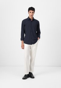 Marineblaue langärmelige Hemdbluse mit spitzem Kragen, kombiniert mit hellbeigen Hosen und schwarzen Schuhen, die ein kontrastierendes Ensemble bilden.
