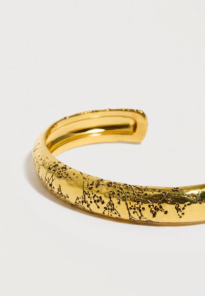 Χρυσό βραχιόλι μανσέτα με υφή, ακανόνιστες μαύρες σημαδούρες πάνω σε μια λεία, γυαλιστερή επιφάνεια, εμφανίζεται σε λευκό φόντο.