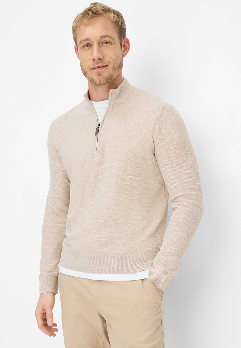 Uomo che indossa un maglione beige con cerniera a tre quarti sopra una camicia bianca e pantaloni beige, in piedi con una mano in tasca, su sfondo bianco.
