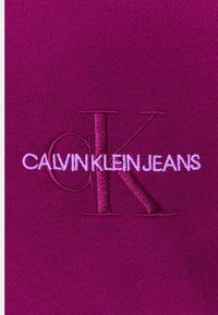 Tessuto viola con loghi ricamati: "CK" in viola scuro e "CALVIN KLEIN JEANS" in viola chiaro, con una texture liscia.