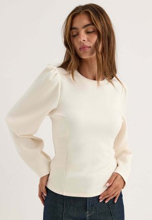 Kvinna med brunt hår som bär en off-white långärmad topp och mörka byxor, poserar med händerna nära höfterna och ögonen slutna.