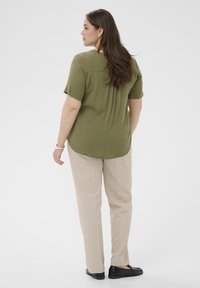 Chemise manches courtes vert olive avec pli dans le dos, associée à un pantalon beige clair et des chaussures noires sans lacets. Texture lisse, coupe décontractée.