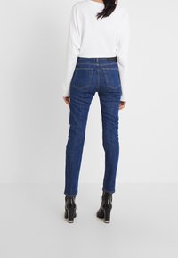 Modré džíny skinny s mírným vzestupem v pase, zadními kapsami a jemným prošíváním, v kombinaci s bílou delší košilí a černými podpatky.