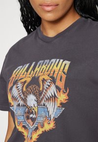 Camiseta de algodón gris con un diseño gráfico que presenta un águila, llamas y el texto "BILLABONG" en letras amarillas y naranjas estilizadas.