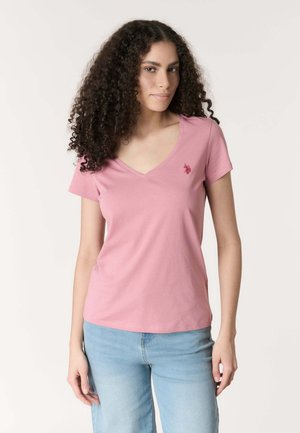 Giovane donna con capelli ricci che indossa una maglietta rosa scollo a V e jeans azzurri chiari, in piedi davanti a uno sfondo chiaro uniforme.