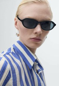 Schwarze ovale Sonnenbrille mit dunklen Gläsern; getragen mit einem blau-weißen gestreiften Kurzarmhemd, das einen klassischen Kragen und eine schlichte Textur aufweist.