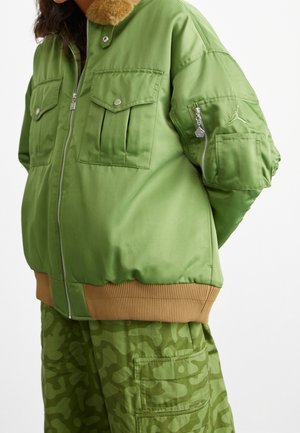 Giacca bomber verde con colletto in pelliccia, chiusura con zip e quattro tasche frontali. Orlo a coste marrone e pantaloni in camouflage verde abbinati.