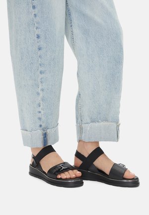 Sandali neri con una fascia larga e fibbia regolabile, caratterizzati da una suola imbottita. Abbinati a jeans azzurri con orli sfrangiati.