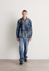 Lichte spijkerblouse met abstracte blauwe patronen, knoopsluiting aan de voorkant en borstzak; gecombineerd met lichtblauwe jeans en zwarte schoenen.