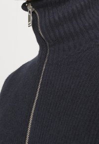 Marineblauer Pullover mit hohem Kragen, gerippter Struktur und einem durchgehenden Reißverschluss mit metallischen Beschlägen an der Naht. Weiches Strickmaterial.