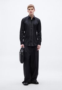 Fekete, hosszú ujjú ing texturált begombolós résszel, laza szabású fekete nadrággal párosítva. A modell egy fekete táskát tart, és lapos cipőt visel.