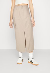 Jupe cargo beige avec poches latérales, fente avant et taille élastique. Associée à des baskets blanches et noires ainsi qu'à des chaussettes blanches.