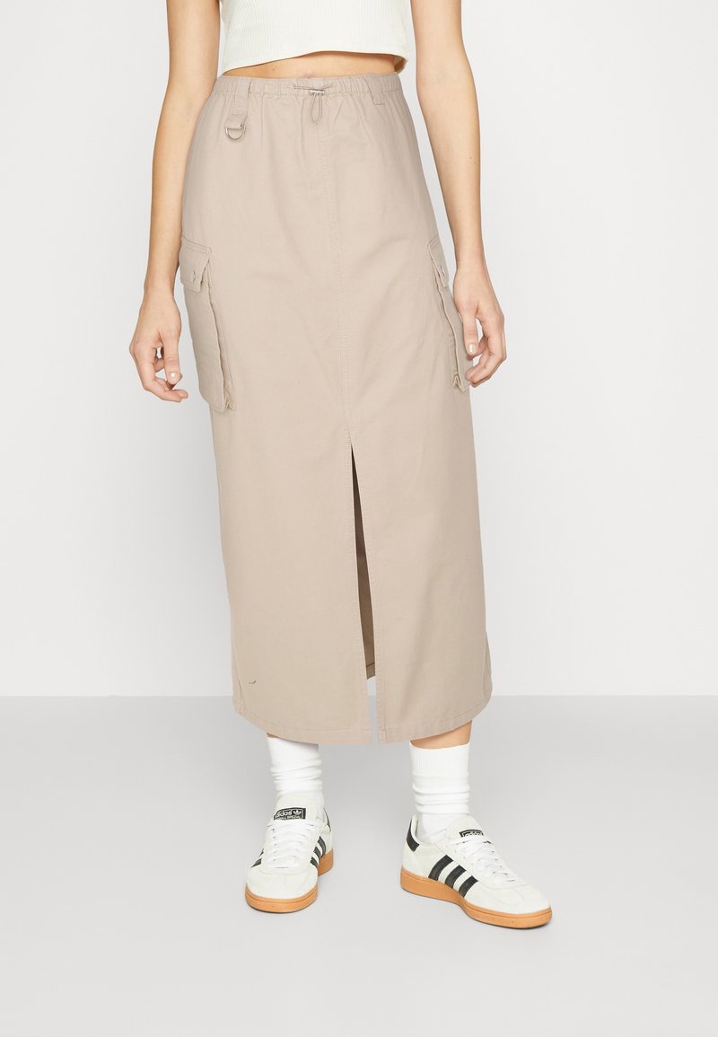 Jupe cargo beige avec poches latérales, fente avant et taille élastique. Associée à des baskets blanches et noires ainsi qu'à des chaussettes blanches.