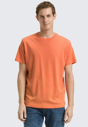 Man with short brown hair wearing plain orange t-shirt and blue jeans, standing against light gray background.