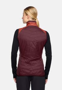 Bordeaux geklede gewatteerde gilet met chevronstiksel, hoge kraag en een slank ontwerp. Heeft contrasterende oranje schouderaccenten en ritszakken aan de zijkanten.