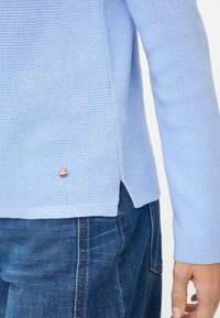Hellblauer Strickpullover mit strukturiertem Muster und Seitenschlitzen, mit einem kleinen Logotag nahe dem Saum, kombiniert mit dunkelblauen Jeans.