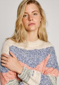 Džemper s više boja, pleten u boji krem, plave i koraljne, s uzorkom u obliku cik-cak i rebrastim manžetama. Materijal izgleda mekano i teksturirano.