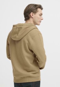 Beige hettegenser med hette, som har en myk tekstur, ribbestrikkede mansjetter og kant, samt en avslappet passform. Bakfra vises sømdetaljer.