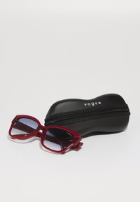 Ochelari de soare roșii cu un design geometric îndrăzneț, având lentile gradient și accesorii din argint, așezați lângă un etui negru cu fermoar etichetat "vogue."