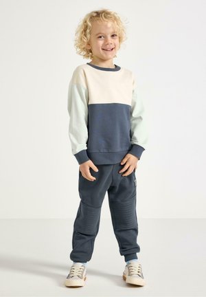 Completo per bambini con felpa color block in crema, blu navy e menta, abbinata a pantaloni scuri. I pantaloni presentano dettagli a coste e tasche laterali.