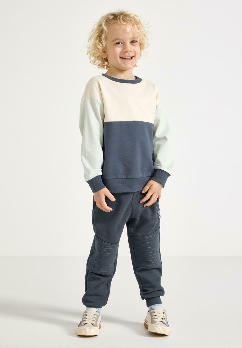 Dětský outfit zahrnující mikinu s barevnými bloky v krémové, námořnické modré a mátové barvě spolu s odpovídajícími tmavými kalhotami. Kalhoty mají žebrované detaily a boční kapsy.