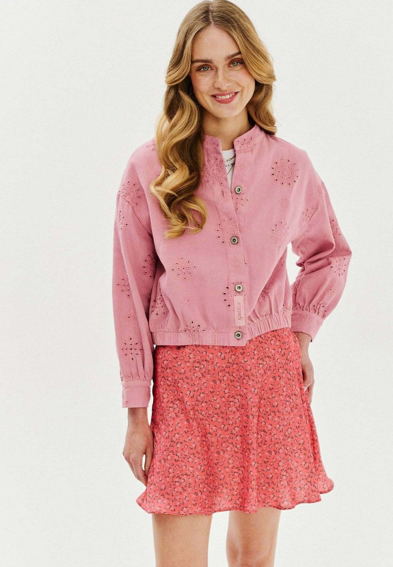 Różowa kurtka z wzorem na wycięcia, zapinana na guziki, z marszczonym dołem, w połączeniu z czerwoną spódnicą w stylu skater z rozwianą sylwetką.