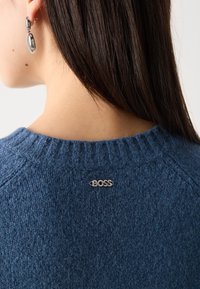 Blauwe gebreide trui met een ronde halslijn en ribbelboord. Voorzien van een zilveren "BOSS"-logo op de achterkant. Zachte textuur en een gemiddeld gewicht.