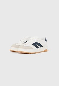 Baltų sportinių batelių pora su smėlio spalvos nosies dangteliais, tamsiai mėlynomis šoninėmis juostelėmis ir storomis šviesiai rudos spalvos padų dalimis, padėta šalia viena kitos ant balto fono.