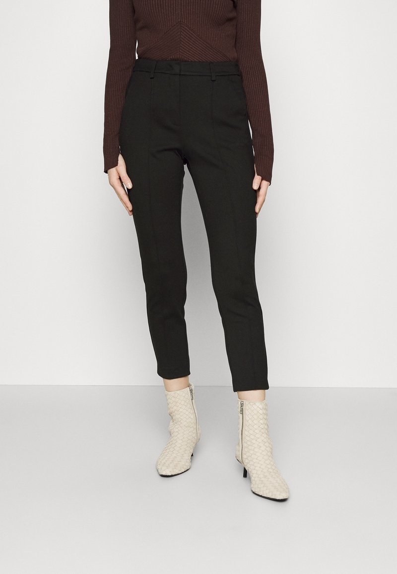 Femme portant un pantalon noir ajusté aux chevilles, un haut côtelé marron foncé à manches longues et des bottines blanches texturées avec fermeture éclair.