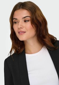Sort jakke med en skarp revers, parret med en ribbet hvid t-shirt. Modellen har langt, brunt bølget hår og et neutralt udtryk.