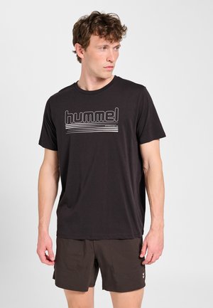 Mężczyzna w czarnym t-shircie marki Hummel i ciemnych szortach, patrzący na bok na jednolite jasne tło.