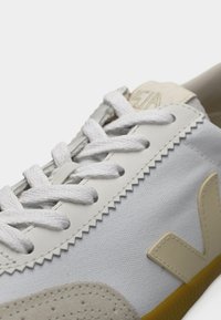 Λευκό καμβαδένιο αθλητικό παπούτσι με μπεζ λεπτομέρειες, υφασμένο λαστιχένιο πέλμα, στρογγυλεμένη μύτη και επίπεδα λευκά κορδόνια. Διαθέτει ραμμένη σχεδίαση και λογότυπο.