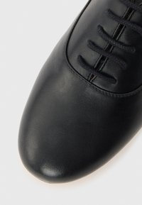 Κοντινό πλάνο μαύρου δερμάτινου παπουτσιού με στρογγυλεμένη μύτη και πέντε σφιχτά δεμένα μαύρα κορδόνια πάνω σε λείο φόντο.