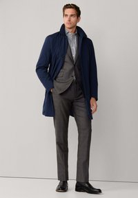 Man in een grijs pak, gestreepte shirt, marineblauwe jas en zwarte nette schoenen staat met één hand in zijn zak tegen een effen achtergrond.