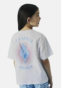 Witte t-shirt met korte mouwen, voorzien van een blauwe schelp graphic met lijnen en de tekst "SEA SHELL HOTEL" in lichtblauwe en roze accenten.