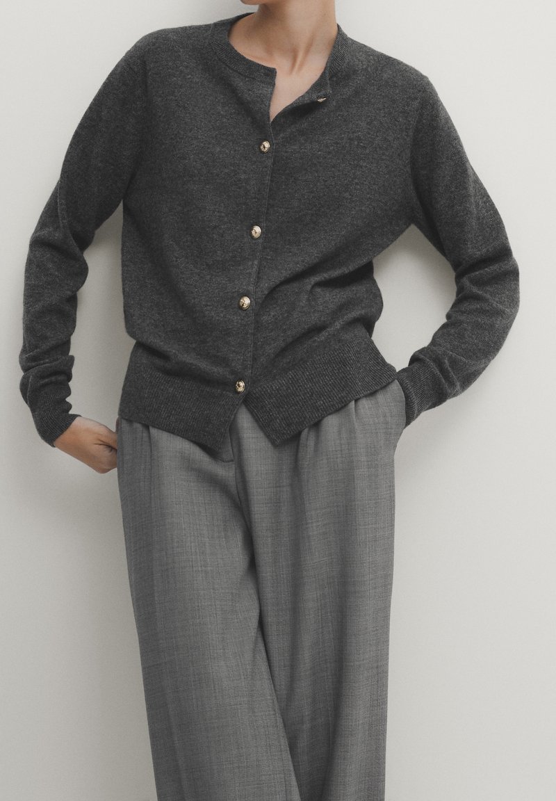 Personne portant un cardigan boutoné gris foncé avec des boutons dorés et un pantalon ample gris clair, se tenant avec les mains partiellement dans les poches.