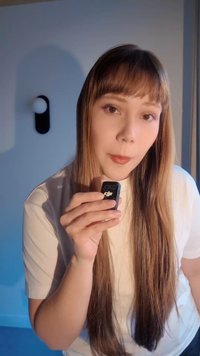 Mladá žena s dlouhými rovnými vlasy drží malé černé zařízení DJI u obličeje a stojí v měkce osvětlené místnosti.