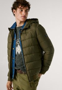 Vadderad olivgrön jacka med avtagbar huva, med quiltad struktur, dragkedjeslutning och två sidofickor, lager på ett mönstrat tröja.