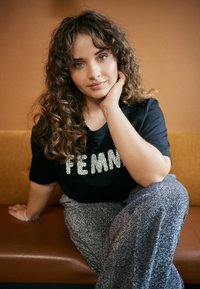 Black t-shirt with sparkly "FEMME" text, paired with silver glittery wide-leg pants. Textured hair and relaxed pose against a warm background.