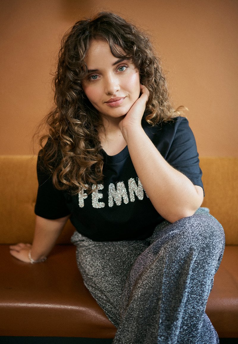 Black t-shirt with sparkly "FEMME" text, paired with silver glittery wide-leg pants. Textured hair and relaxed pose against a warm background.