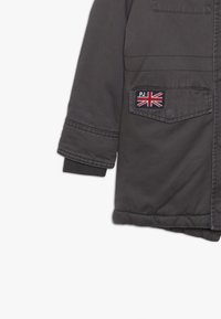 Ciemnoszara kurtka o miękkiej fakturze, z zamkiem błyskawicznym, bocznymi kieszeniami oraz naszywką z flagą Wielkiej Brytanii na lewej piersi.