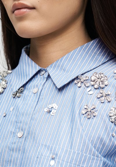Κοντινό πλάνο λαιμού και κάτω μέρους προσώπου που φοράει μπλε και λευκό ριγέ πουκάμισο με διακοσμήσεις από παγιέτες και χάντρες σε μορφή λουλουδιού στον ώμο και το στήθος.