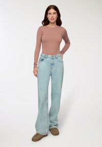 Różowa bluzka z długim rękawem w odcieniu stonowanym, zestawiona z jasnoniebieskimi spodniami typu wide-leg o wysokim stanie. Miękka tekstura, swobodny krój, z brązowym obuwiem.