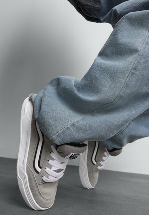 Szare zamszowe sneakersy z białymi gumowymi podeszwami i czarnymi akcentami, w zestawieniu z jasnoniebieskimi dżinsami, skupione na detalach obuwia.