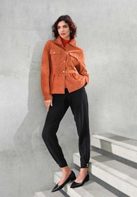 Suède oranje jasje met twee voorkruimelzakken en gouden knopen, gecombineerd met zwarte tapered broek en zwarte puntige hakken, op een betonnen trap.