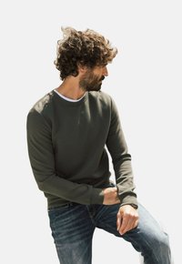 Sudadera verde oliva con cuello redondo y puños de canalé, combinada con jeans claros desgastados. Diseño sencillo y moderno.